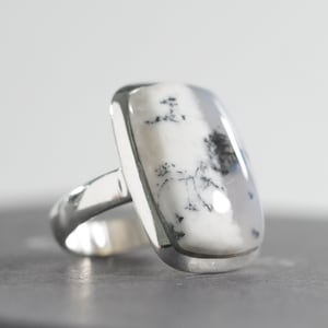 May include: A silver ring with a rectangular stone displaying a dendritic pattern. The stone is mainly white with black and grey inclusions, set in a polished silver band. The ring has a classic, elegant design.
