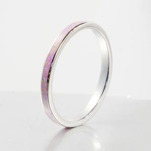 May include: A thin, polished silver-tone metal ring featuring a smooth, inlaid band of iridescent pink material. The inlay displays subtle flecks of color throughout its surface, set within a simple, rounded metallic frame with a reflective finish.