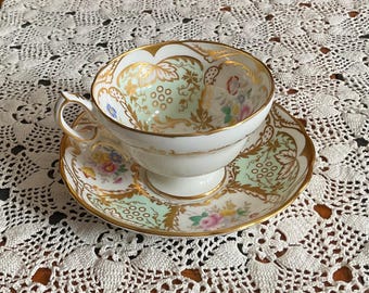 May include: A vintage teacup and saucer set. The white porcelain cup and saucer feature a gold trim and a delicate floral and vine design with a light green background. The set is placed on a white lace tablecloth.