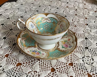 May include: A vintage teacup and saucer set. The cup and saucer are white with gold trim and feature a teal and gold floral design. The saucer has a matching design with colorful flowers. The set is placed on a white lace tablecloth.