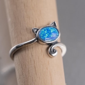 May include: A silver ring featuring a cat-shaped design with two small pointed ears and a curled tail. The center of the cat head is set with an oval-shaped blue opal stone that displays iridescent flecks of light blue and green throughout the surface.