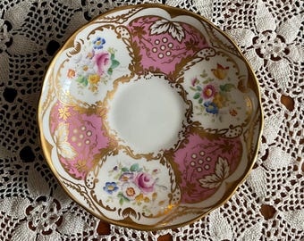 May include: A vintage porcelain saucer with a white center and a pink and gold floral design. The saucer features a scalloped edge with gold trim and is decorated with colorful flowers and leaves. The overall design is ornate and elegant.