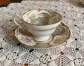 May include: A white teacup and saucer set with gold trim and floral and leaf designs. The teacup has a handle and sits on a matching saucer. The set is placed on a white lace tablecloth, creating an elegant aesthetic.