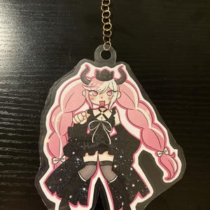 Puede incluir: Un llavero de acrílico transparente con un personaje de anime de estilo rosa y negro con cuernos y coletas, que lleva un vestido negro con estampado de galaxia.