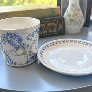 May include: A white ceramic cup and saucer set. The cup features a blue and green illustration of a person with floral details. The saucer has a white center with a blue and white patterned rim featuring small repeating geometric shapes.