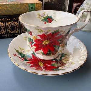 May include: A white porcelain teacup and saucer set featuring a vibrant red poinsettia, green holly leaves, red berries, and pinecone illustrations. The teacup has a scalloped rim and a gold-trimmed edge, with a matching floral pattern inside.