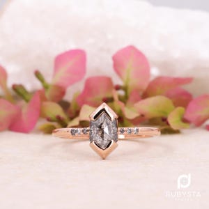 May include: Rose gold ring featuring a gray, hexagonal-cut diamond center stone. The ring has a geometric design with small accent stones along the band. Pink flowers and a white background are in the background.