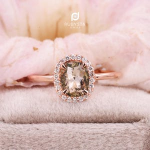 Peut inclure: Une bague en or rose avec une pierre centrale ovale de couleur gris-brun moucheté, maintenue par quatre griffes. La pierre centrale est entourée d'un halo de petites pierres blanches rondes et claires. La marque RUBYSTA JEWELS est visible au-dessus.