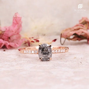 May include: Rose gold engagement ring with a large oval-cut, salt and pepper diamond center stone. The band is adorned with small, sparkling accent diamonds. The ring is set against a soft, neutral background with pink floral accents.
