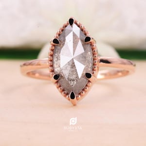May include: Rose gold ring featuring a marquise-cut, gray diamond. The diamond is set in a beaded bezel with black accents. The ring's band is a simple, polished design. The ring is displayed on a light-colored surface.