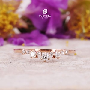 May include: Rose gold ring with a curved design, adorned with multiple clear, sparkling gemstones. The ring is set against a soft, neutral background with blurred purple flowers and the text "RUBYSTA JEWELS".