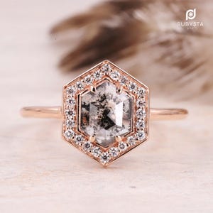 Peut inclure: Une bague en or rose avec un diamant hexagonal poivre et sel au centre, entouré d'un halo de petits diamants blancs ronds. Le sertissage est de forme hexagonale et l'anneau est un fil d'or rose lisse. Le logo RUBYSTA USA est visible.