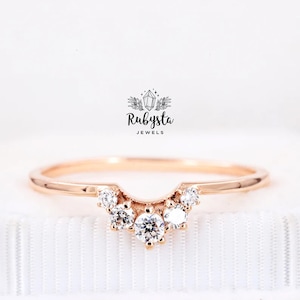 May include: Rose gold ring with a curved design, featuring several small, clear gemstones. The ring is set against a white background. The brand name "Rubysta Jewels" is visible in the image.
