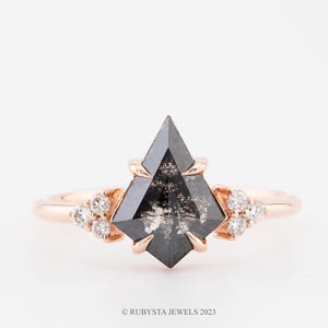 Op de afbeelding: Een roségouden ring met een vliegervormige zout-en-peper diamant met een gespikkelde grijze en zwarte uitstraling. De ringband is aan beide zijden geaccentueerd met een cluster van drie kleine ronde witte diamanten in een klauwzetting.