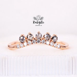 May include: A delicate rose gold ring with a curved band and a row of small diamonds. Five larger, grey diamonds are set in the center of the band, creating a crown-like design. The ring is displayed on a white background with the text "Rubysta Jewels".