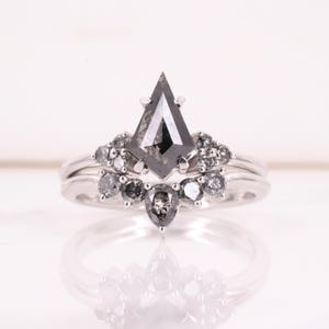 Peut inclure: Une bague en argent ornée d'un diamant gris central en forme de cerf-volant, entouré de petits diamants gris ronds et en forme de poire disposés en grappe incurvée le long de l'anneau, créant un aspect superposé et symétrique.