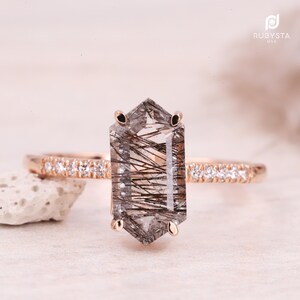 May include: A rose gold ring featuring a large, clear, hexagonal gemstone with black inclusions. The band is adorned with small, clear accent stones. The ring is set on a neutral surface.