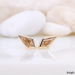 May include: A pair of gold-colored stud earrings. Each earring features a geometric, kite-shaped design with a light brown gemstone center. The earrings are displayed on a reflective surface, with a blurred white background.