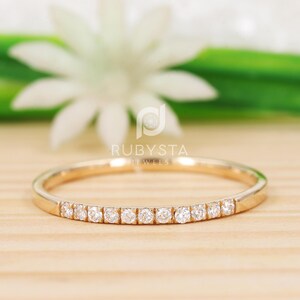 May include: A gold band ring with a row of small, clear gemstones. The ring is set against a wooden surface with a blurred white flower and green foliage in the background. The brand name "RUBYSTA JEWELS" is visible.