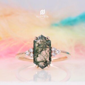 Peut inclure: Une bague en or rose avec une pierre centrale en agate mousse hexagonale allongée présentant des inclusions dendritiques vert foncé. La pierre est fixée par six griffes, flanquée de chaque côté par trois petits diamants blancs ronds.