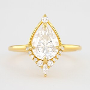 May include: A pear-shaped diamond engagement ring set in yellow gold with a halo of smaller diamonds and a delicate, pointed design.