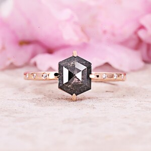 May include: A rose gold ring featuring a large, hexagonal, dark gray diamond as the centerpiece. The band is adorned with small, round diamonds. The background is a soft pink, creating a contrast with the ring's design.