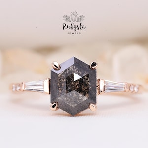 May include: A rose gold engagement ring with a hexagonal black diamond and two baguette diamonds on the band. The ring is set on a white background.