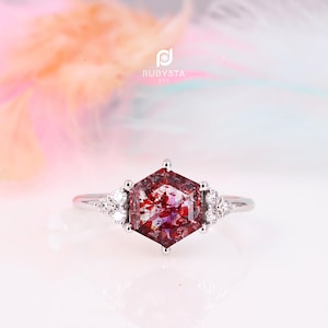 May include: A silver-toned ring featuring a central hexagonal-cut gemstone with deep red and purple inclusions. The center stone is flanked by two clusters of three small, clear round-cut stones on each side. The text RUBYSTA USA is visible.