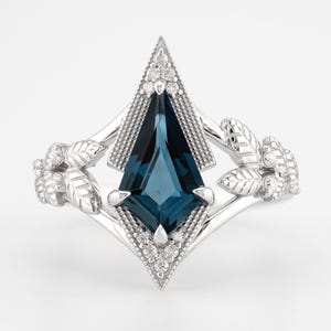 May include: A silver-toned ring featuring a central kite-shaped dark blue gemstone surrounded by small clear stones. The band is designed with metallic leaf motifs on both sides, and the setting includes intricate milgrain detailing around the center.