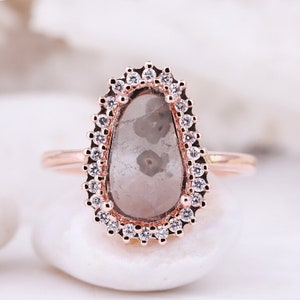 Peut inclure: Une bague en or rose ornée d'une pierre grise translucide en forme de poire avec des inclusions visibles au centre. La pierre est entourée d'un halo de petites pierres blanches rondes et claires sur une monture festonnée en or rose.