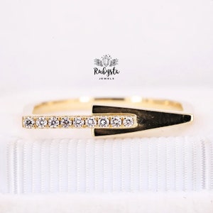 May include: A gold-colored ring with a geometric design. The ring features a row of small, clear gemstones set in a straight line. The ring has a unique arrow-like shape. The brand name "Rubysta Jewels" is visible in the image.