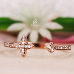 May include: Rose gold open ring featuring a four-leaf clover design on one end and a diamond-shaped accent on the other. The band is encrusted with small, sparkling stones. The ring is set against a soft, blurred background.