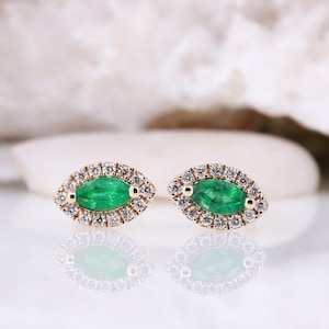 May include: A pair of gold stud earrings with a marquise-cut emerald center stone, surrounded by a halo of small, sparkling diamonds. The earrings are displayed on a reflective surface, showcasing their intricate design and vibrant green color.