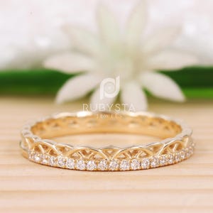 May include: A gold-colored ring with a delicate design. The ring features a row of small, clear gemstones set along the band. The band has an intricate, openwork pattern. The brand name "RUBYSTA JEWELS" is visible in the background.