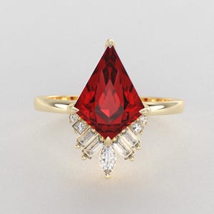 May include: A gold ring featuring a large kite-shaped red gemstone as the centrepiece. The setting is accented with a cluster of smaller clear baguette and marquise-cut stones arranged in a fan-like pattern beneath the main red gem.