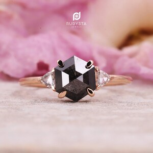 May include: A rose gold ring featuring a large, hexagonal black diamond center stone, flanked by two smaller, triangular white diamonds. The ring has a delicate band and is set against a soft pink background. The brand name "RUBYSTA JEWELS" is visible.