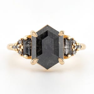 May include: A gold ring with a large, hexagonal, black diamond in the center. The diamond is surrounded by smaller, black diamonds set in a gold band.