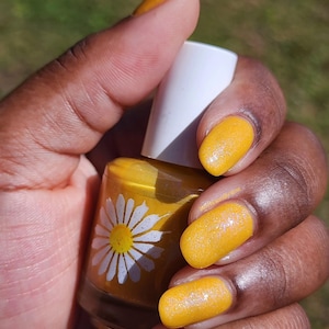 May include: A hand holding a bottle of yellow nail polish with a white daisy design. The nails are painted a matching yellow color. The background is blurred green.