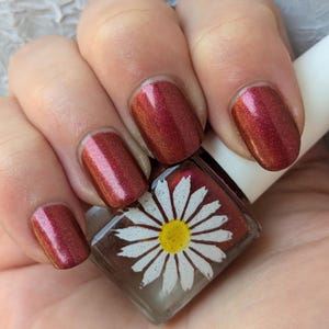 May include: A clear glass bottle of red nail polish with a white daisy on the label. The bottle is being held by a hand with red nail polish on the nails.