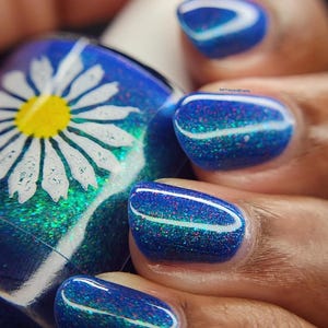 May include: Close-up of a hand with manicured nails. The nails are painted a deep blue with a shimmering, glittery effect and a white stripe. A nail polish bottle with a daisy design is visible.