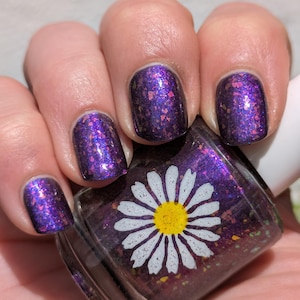 May include: A glass bottle of nail polish with a white daisy graphic on the front. The polish is a deep purple base filled with iridescent, multi-colored flakes. The same purple, flake-filled polish is applied to the fingernails, creating a matching look.