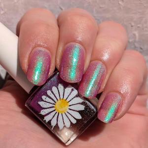 May include: Close-up of a hand with five fingernails painted with a pink and green iridescent nail polish. The nail polish bottle is in the foreground and has a white daisy design on the label.