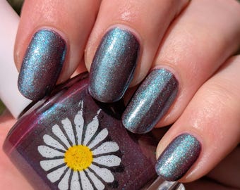 May include: Close-up of a hand with polished nails and a nail polish bottle. The nails are painted with a shimmering teal and purple polish. The nail polish bottle is dark purple with a white daisy design.
