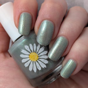 May include: A hand holding a bottle of nail polish with a daisy design. The nails are painted a shimmering, light green color. The nail polish bottle is gray with a white daisy and yellow center.