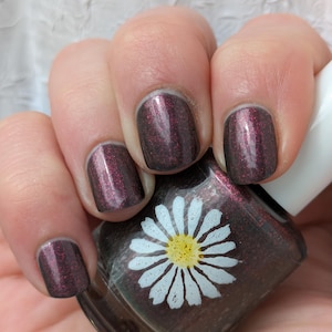 May include: A glass bottle of dark plum nail polish with a shimmering red glitter finish. The bottle features a white daisy graphic with a yellow center printed on the front. The nail polish color on the fingernails matches the bottle.