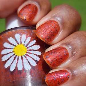 May include: A glass bottle of nail polish featuring a white daisy graphic with a yellow center. The polish is a vibrant orange hue with iridescent blue and purple flakies, matching the color and texture applied to the fingernails shown nearby.