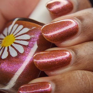 May include: Close-up of a hand with polished nails. The nails are painted with a shimmering, reddish-brown nail polish. The polish has a glittery effect, reflecting light. A bottle of nail polish with a daisy design is visible.