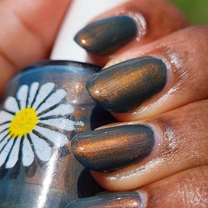 May include: Close-up of a hand with polished nails in a dark teal color with a copper shimmer. A nail polish bottle with a white cap and a daisy design is in the background. The nail polish has the text "berriezandkreme".