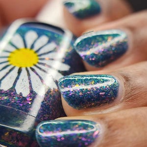 May include: A glass bottle of nail polish featuring a white daisy graphic with a yellow center against a dark teal base filled with iridescent, multi-colored glitter flakes. The polish is applied to fingernails, showing a matching dark teal finish.