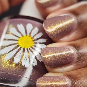 May include: A glass bottle of nail polish featuring a white daisy graphic with a yellow center, held alongside fingernails painted in a mauve-toned polish with a fine gold shimmer finish that reflects light across the surface of each nail.